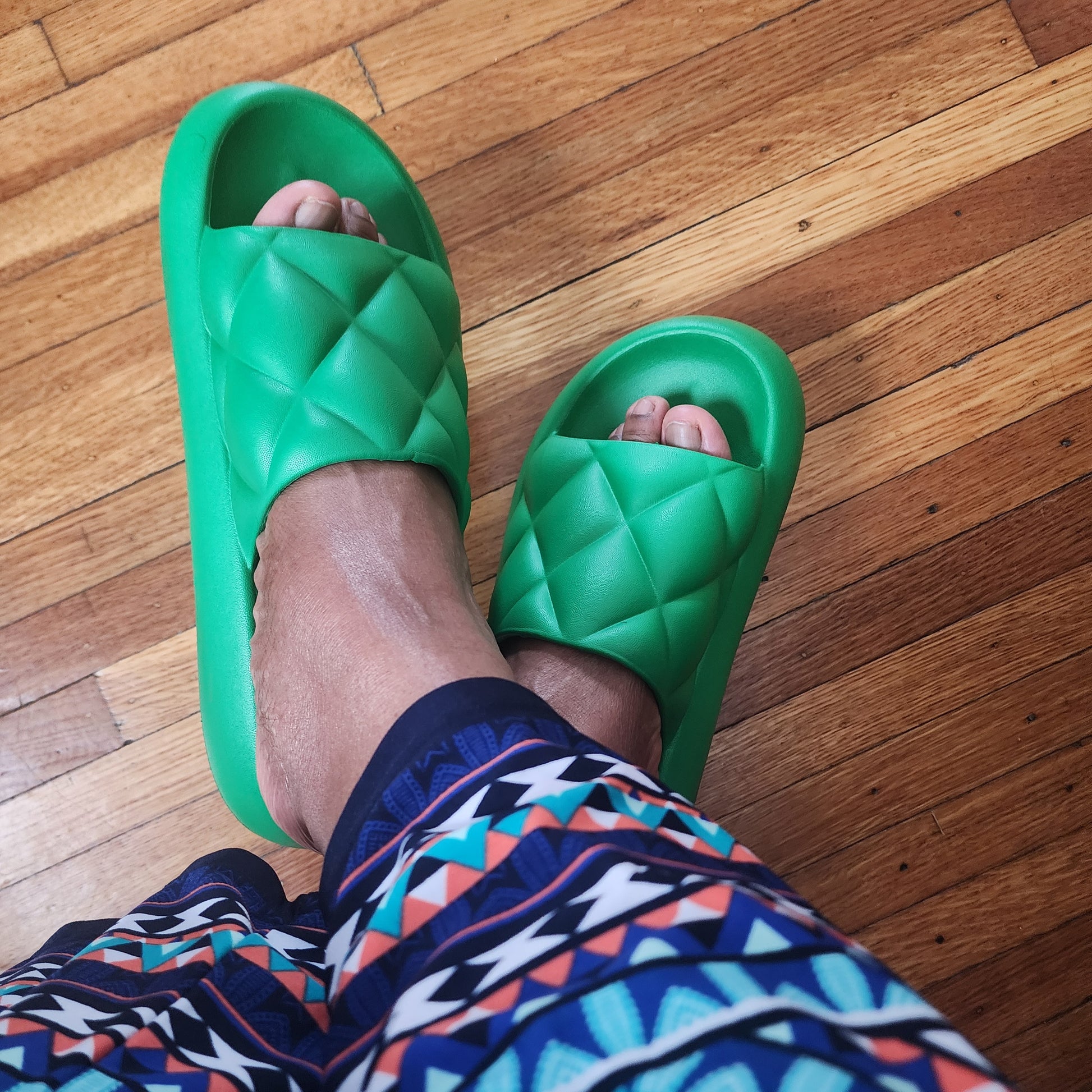 Green slides on feet with patterned pants on a wooden floor
